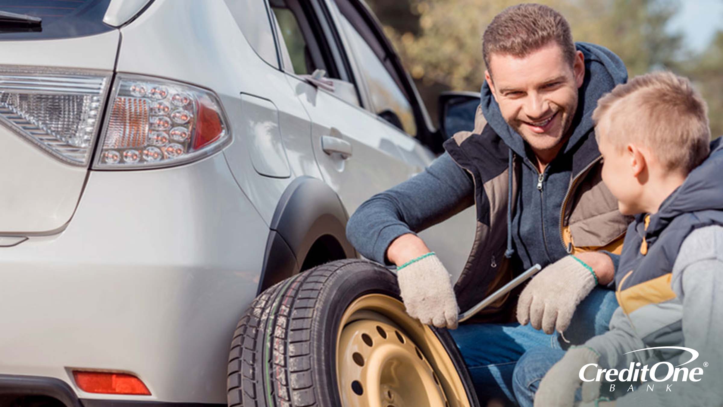 A man smiles at his young son as they crouch beside their car, having just removed the wheel to change a flat tire. This is a common auto repair emergency.